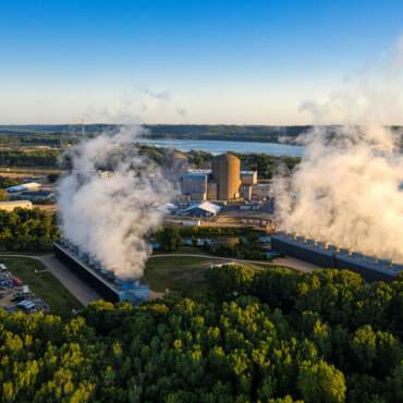 Panoramica aerea di un grande impianto industriale, una centrale nucleare con torri di raffreddamento attive, situata in un'area verde vicino a un fiume, a simboleggiare l'importanza della gestione delle emissioni industriali.